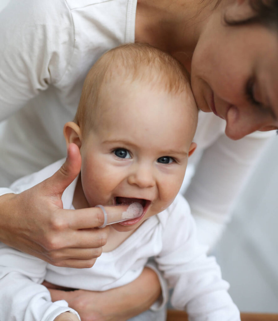 Lachendes Baby mit ersten Zähnchen, Mutter berührt zärtlich das Kinn beim innigen Familienmoment.