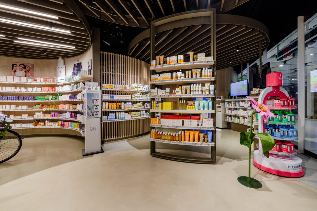 Moderne Apotheke mit geschwungenen Regalen, stilvollem Holzdecken-Design und auffälligem rosa Blickfang im Innenraum.