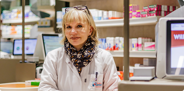 Freundliche Apothekerin im weißen Kittel berät Kunden hinter dem Tresen einer modernen Apotheke.