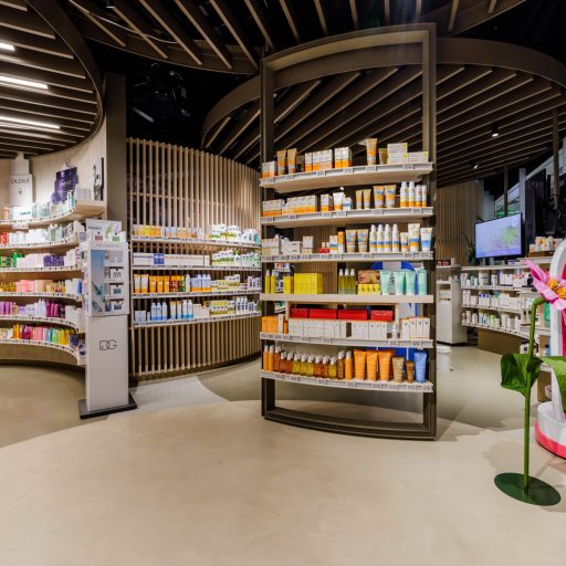 Moderne Apotheke mit geschwungenen Regalen, stilvollem Holzdecken-Design und auffälligem rosa Blickfang im Innenraum.